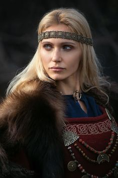 a woman with long blonde hair wearing a fur collar and beaded headband, looking at the camera