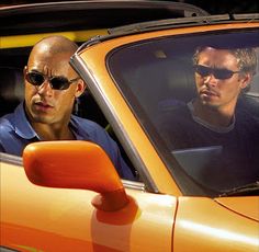 two men sitting in the drivers seat of an orange sports car, one with sunglasses on