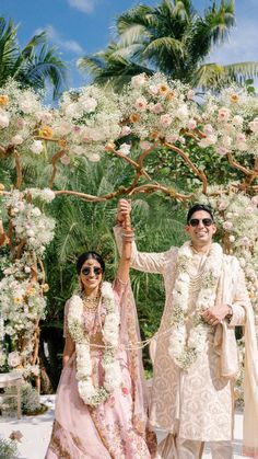 two people standing in front of a flower covered arbor with flowers all around them and one person holding the other hand up