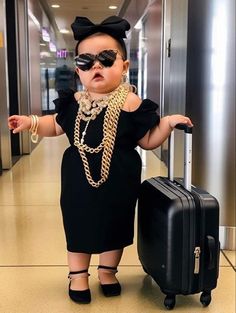 a baby dressed up as a woman with a suitcase and chain around her neck, standing in an airport hallway