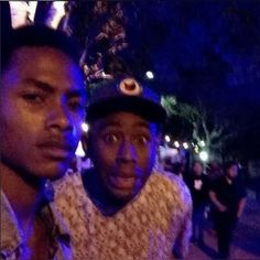 two young men standing next to each other on a street at night with people in the background