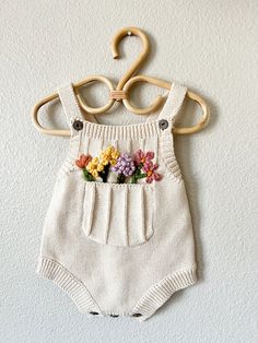 a white knitted romper with flowers on it hanging from a wooden hanger