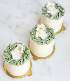 two white cakes decorated with green leaves and flowers on top of a marble countertop