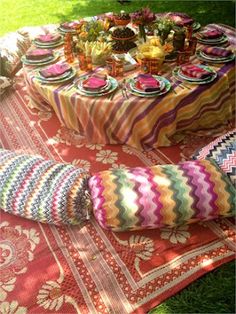 a colorful table cloth with many plates and cups on it sitting in the grass next to a picnic blanket