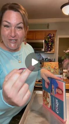 a woman holding up a book in her hand while standing next to a kitchen counter