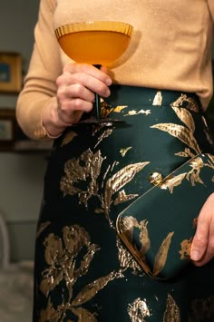 a woman holding a drink in her hand while wearing a gold and green skirt with flowers on it