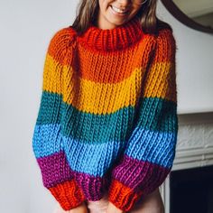 a woman wearing a multicolored sweater standing in front of a fireplace with her hands on her hips