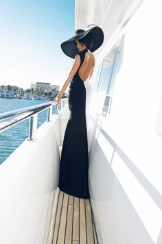 a woman in a long black dress and hat standing on the side of a boat