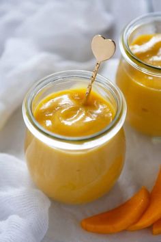 two jars filled with yellow liquid next to carrots on a white cloth and a wooden spoon