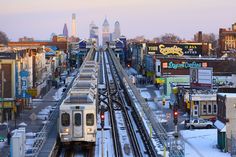 a train is traveling down the tracks in front of some buildings and skyscrapers on a snowy day