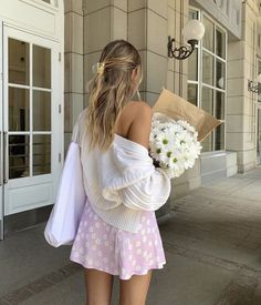 a woman in a white sweater and pink skirt holding flowers walking down the street with her back to the camera