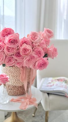 pink roses in a basket sitting on a table next to a white chair and window