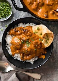 two black bowls filled with rice and meat curry next to silver spoons on a gray surface