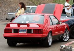 the back end of a red sports car parked in a parking lot with people looking at it