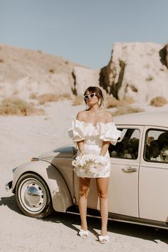 a woman standing next to a white car in the desert wearing sunglasses and a dress with flowers on it