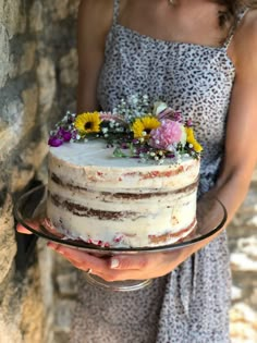 a woman holding a cake with flowers on it in front of a stone wall and wearing a dress