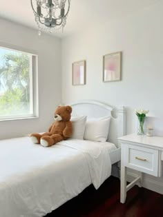 a teddy bear sitting on top of a white bed in a bedroom next to a window