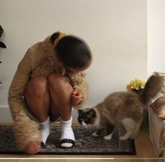 a woman kneeling down next to a cat on a rug with her legs crossed,