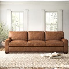 a brown leather couch sitting on top of a wooden floor next to a rug and windows