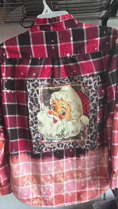 a red and black checkered shirt with a santa clause on the chest, in front of a hanger