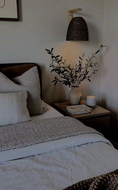 a bed with white sheets and pillows next to a table with a vase on it