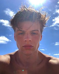 a man with no shirt on standing in front of blue sky and clouds, looking at the camera