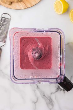 a blender filled with pink smoothie next to sliced lemons and grater