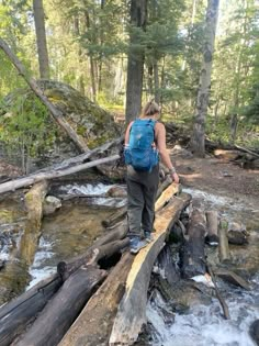 a woman walking across a fallen log over a river in the woods on a sunny day