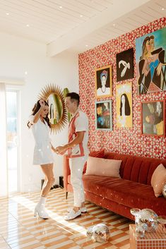 a man and woman standing next to each other in a living room with pictures on the wall