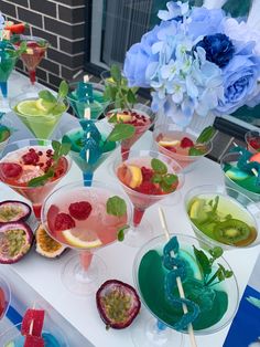 there are many drinks on the table with fruit in glasses next to flowers and blue hydrangeas