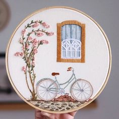 a person holding up a small embroidery kit with a bicycle and flowers in front of a window