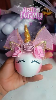 a hand holding a small white ball with a pink unicorn face and gold horn on it