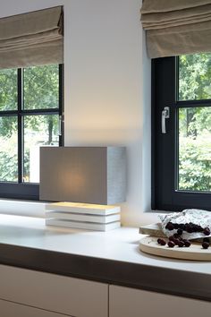 a table with a lamp, plate and fruit on it in front of two windows