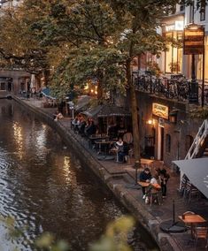 people sitting at tables on the side of a river in front of buildings and trees