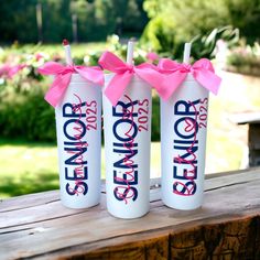 three white water bottles with pink bows are sitting on a wooden bench in front of some flowers