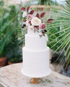 a white wedding cake with pink flowers on top and greenery around the edges is sitting on a wooden table