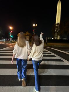 two girls walking across a crosswalk in front of the washington monument, at night