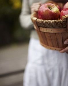 a woman holding a basket full of apples in her hands while wearing a white dress