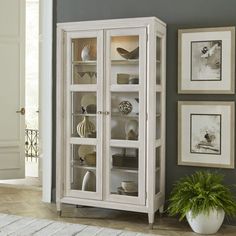 a white china cabinet with glass doors and shelves in the corner next to a potted plant