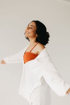 a woman in an orange top and white blazer smiles as she stands with her arms outstretched