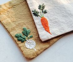 two towels with embroidered carrots on them are sitting next to each other, one is white and the other has green leaves