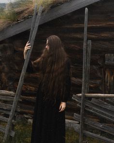 a woman with long hair standing in front of a log cabin and holding a stick