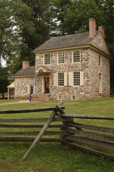 an old stone house with a split rail fence in front of it and trees around the yard