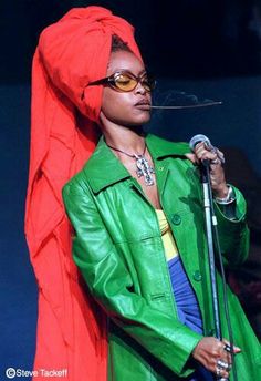 a woman in a green jacket holding a microphone and wearing sunglasses with an orange scarf on her head