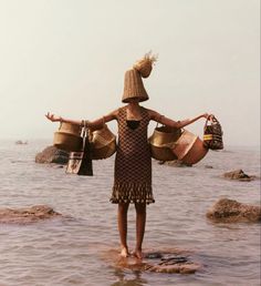 a woman standing in the water with two baskets on her head and one hand outstretched