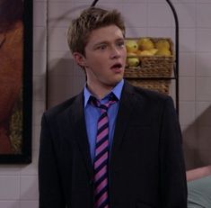 the young man is dressed in a suit and pink striped tie, standing next to a basket of fruit