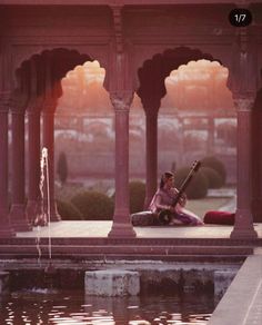 a woman sitting on the ground next to a body of water with an instrument in her hand