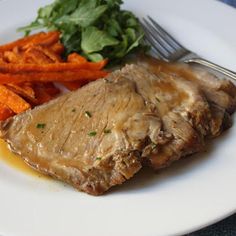 a white plate topped with meat and carrots next to a fork on a blue table cloth