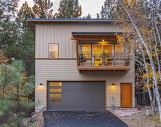 a two story house in the woods with a large garage and attached deck above it