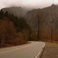 an empty road in the mountains with trees on both sides and foggy skies above